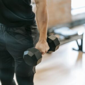 Unrecognizable muscular male in black sportswear lifting heavy dumbbell during weightlifting workout in light gym with bench on blurred background