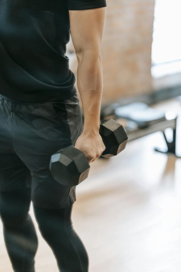 Unrecognizable muscular male in black sportswear lifting heavy dumbbell during weightlifting workout in light gym with bench on blurred background